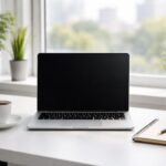 Mesa de escritório moderna com laptop, xícara de café, caderno e caneta, com janela ao fundo mostrando plantas e vista da cidade.